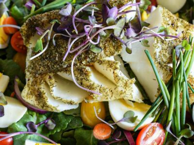 A close up shot of a large serving bowl filled with baby kale salad, with chopped hard boiled eggs, cherry tomatoes, chives, microgreens and baked cod fish covered in dill over the top