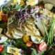 A close up shot of a large serving bowl filled with baby kale salad, with chopped hard boiled eggs, cherry tomatoes, chives, microgreens and baked cod fish covered in dill over the top