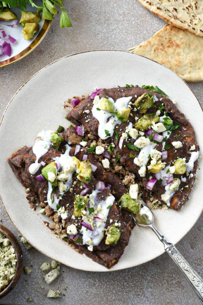 An overhead shot of a white plate with a fork under two enfrijoladas pita pockets filled with ground turkey and topped with a black bean sauce, red onion, feta, avocado and cilantro