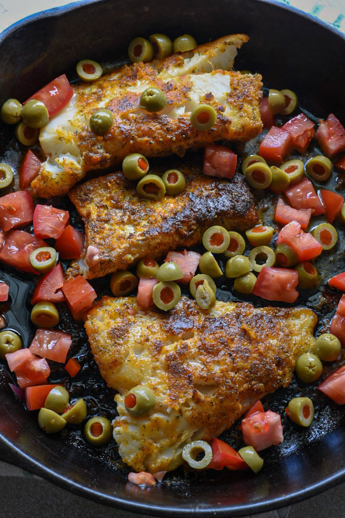 A cast iron skillet filled with three golden white fish fillets, green olives and chopped tomatoes