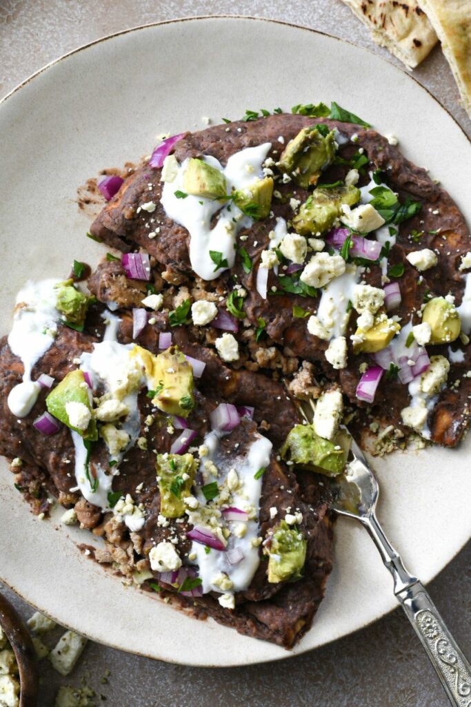 A close overhead shot of a white plate with a fork under two enfrijoladas pita pockets filled with ground turkey and topped with a black bean sauce, red onion, feta, avocado and cilantro