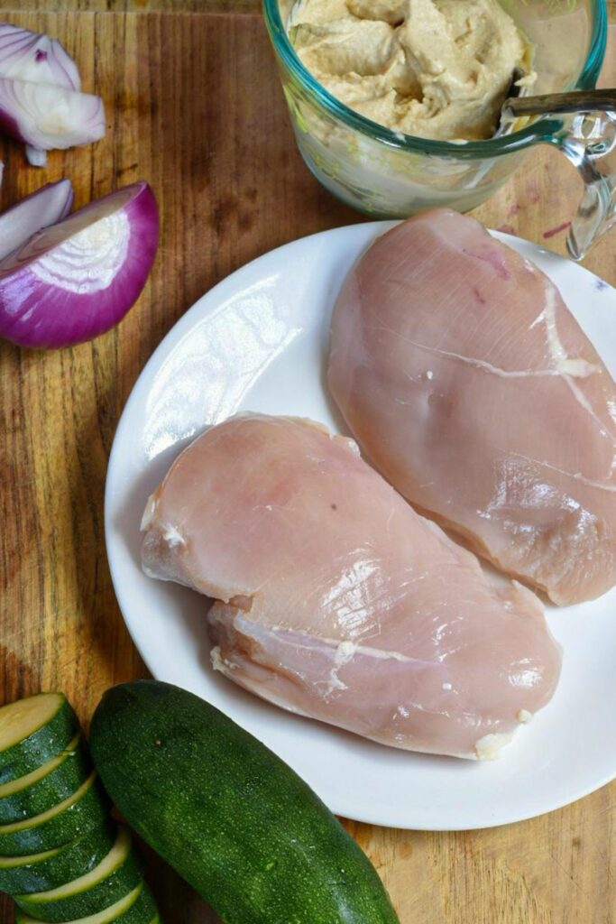 An overhead shot of two large raw chicken breasts on a plate, with sliced zucchini, red onion and hummus in the shot