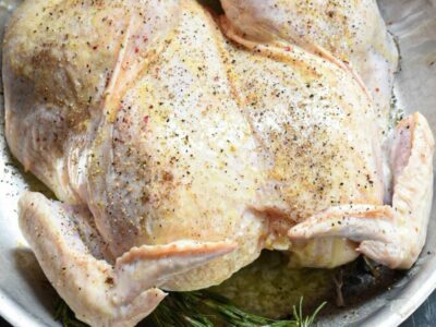 A spatchcocked whole chicken in a stainless steel skillet with rosemary under it