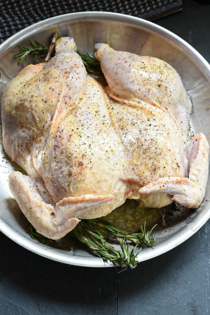 A spatchcocked whole chicken in a stainless steel skillet with rosemary under it