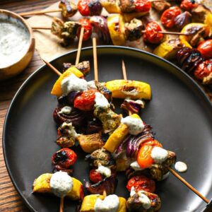A black plate with 4 kabobs on top filled with bright grilled vegetables and lemon dill chicken, with a greek yogurt sauce drizzled over the top