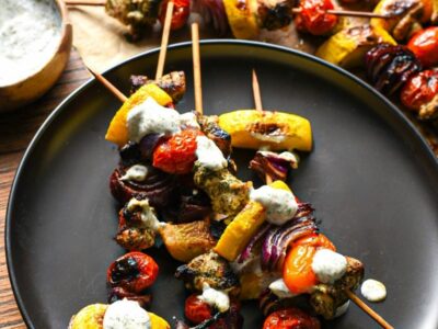 A black plate with 4 kabobs on top filled with bright grilled vegetables and lemon dill chicken, with a greek yogurt sauce drizzled over the top