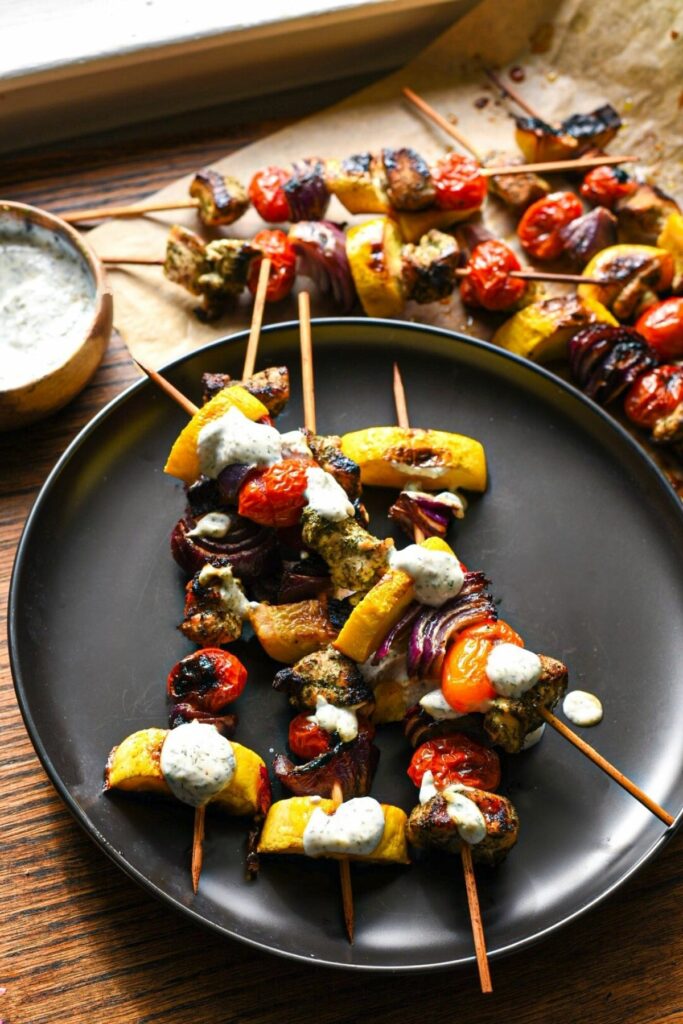 A black plate with 4 kabobs on top filled with bright grilled vegetables and lemon dill chicken, with a greek yogurt sauce drizzled over the top