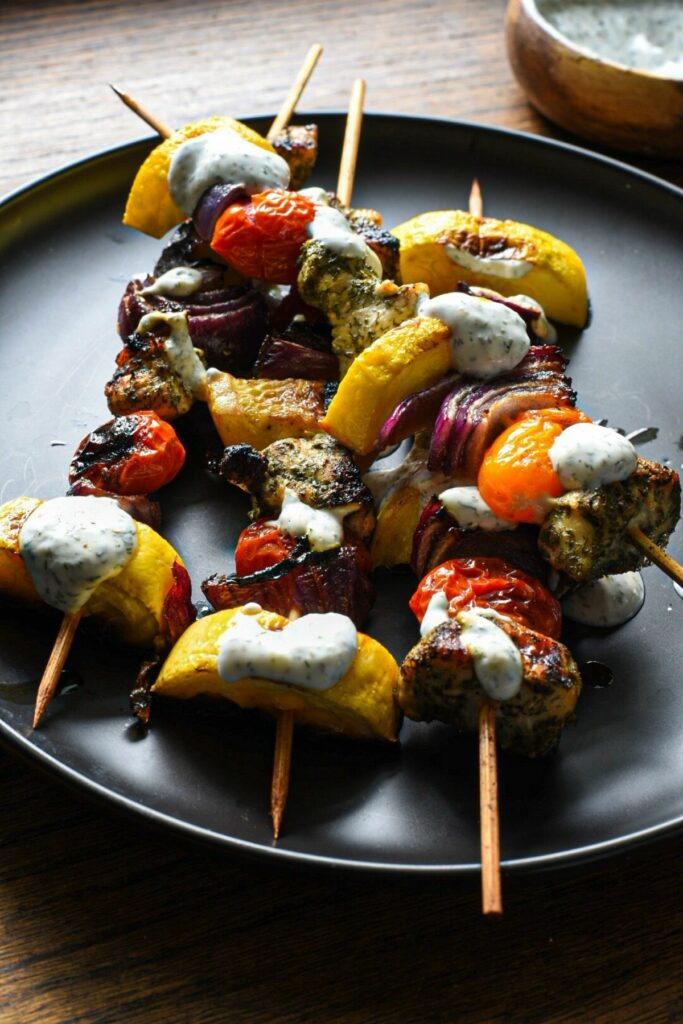A close shot of 4 chicken and vegetable kabobs with a lemon dill greek yogurt sauce drizzled over the top on a black plate with a wooden surface underneath 