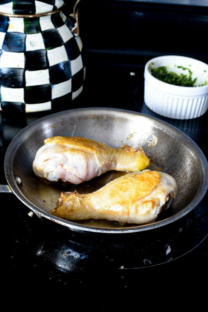 a small stainless steel skillet on an electric stove filled with two cooking chicken drumsticks that are golden brown