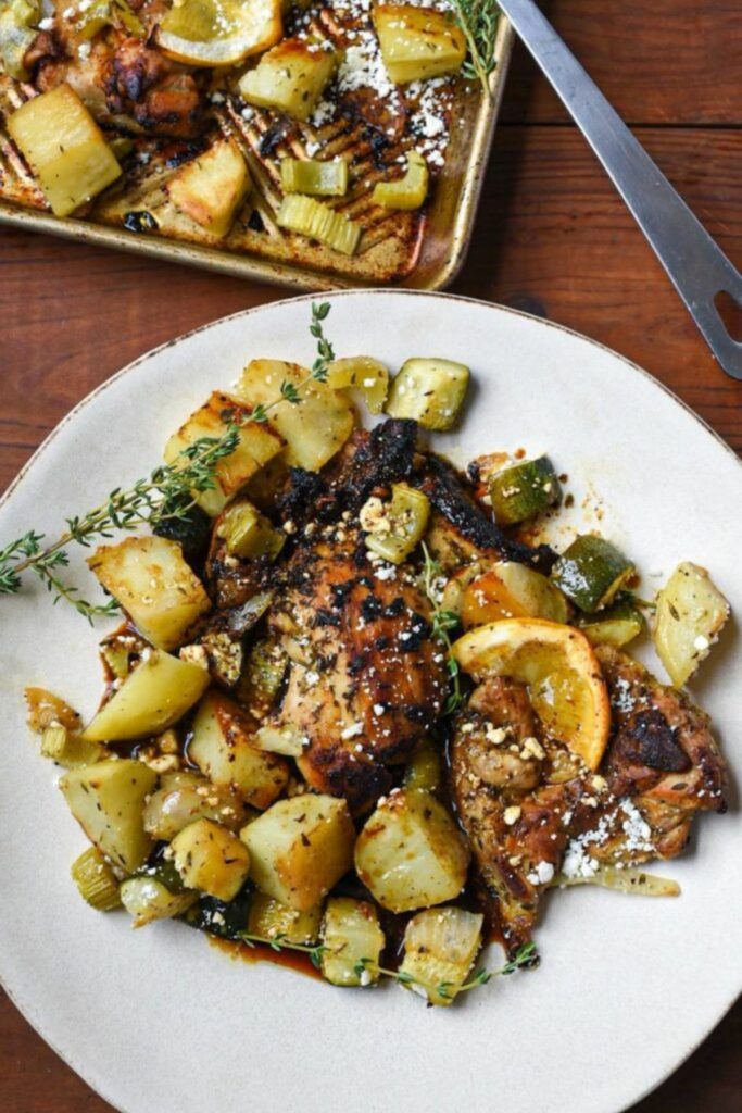 A plate filled with cooked chicken thighs and potatoes, with thyme and lemon around it and a sheet pan with the same ingredients in the corner of the image