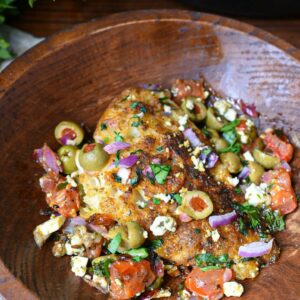 A close up image of mediterranean baked cod recipe, with a golden baked white fish filet topped with green olives, red onion, parsley and tomatoes