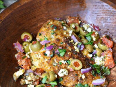 A close up image of mediterranean baked cod recipe, with a golden baked white fish filet topped with green olives, red onion, parsley and tomatoes