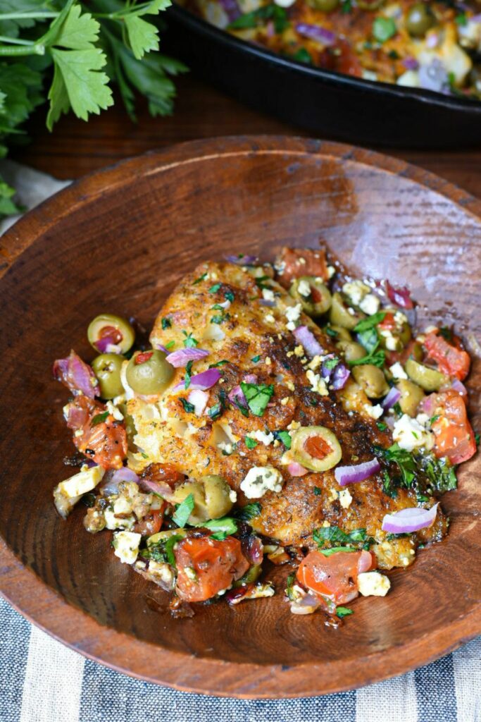 A close up image of mediterranean baked cod recipe, with a golden baked white fish filet topped with green olives, red onion, parsley and tomatoes