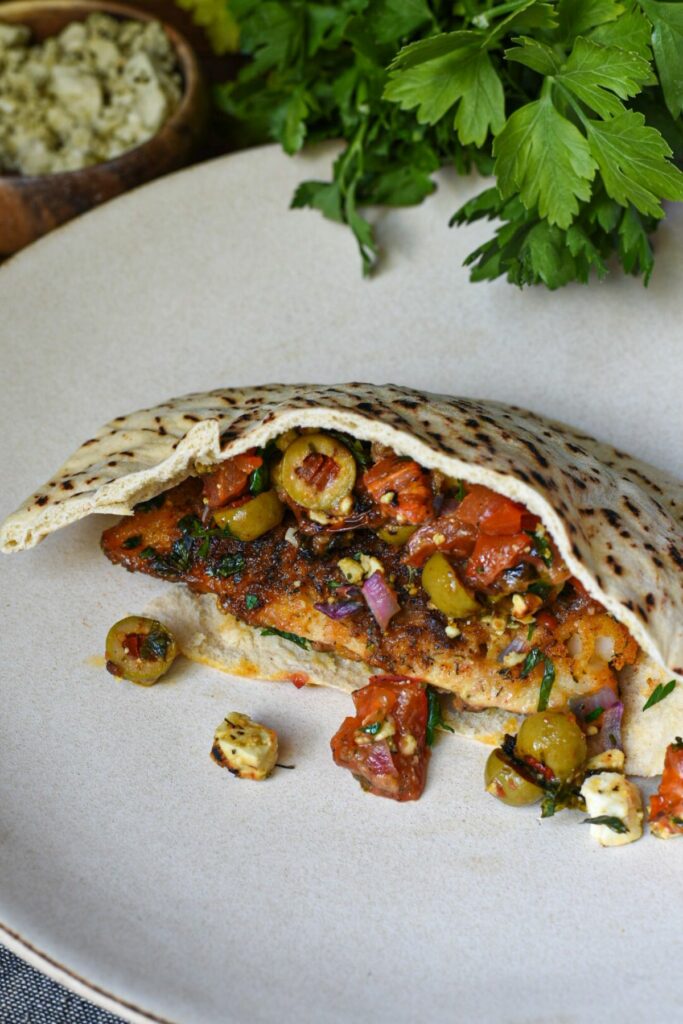 A pita pocket filled with a baked white fish fillet, sliced green olives, chopped tomatoes, red onion, parsley and feta.