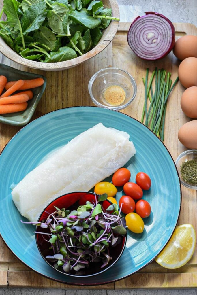An overhead ingredients shot including a blue plate filled with raw cod loin, cherry tomatoes, a red bowl of microgreens, a wooden bowl filled with baby kale, a chopped red onion, baby carrots, 4 hard boiled eggs, chives, lemon and spices