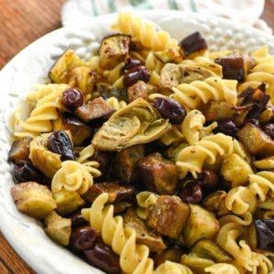 A large white porcelain serving bowl filled with fusilli pasta and roasted eggplant, olives and artichoke hearts