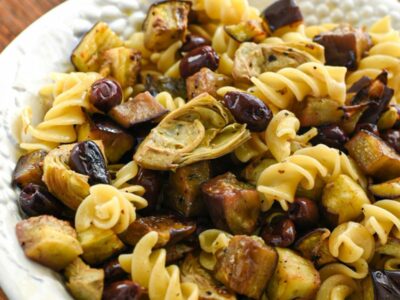 A large white porcelain serving bowl filled with fusilli pasta and roasted eggplant, olives and artichoke hearts