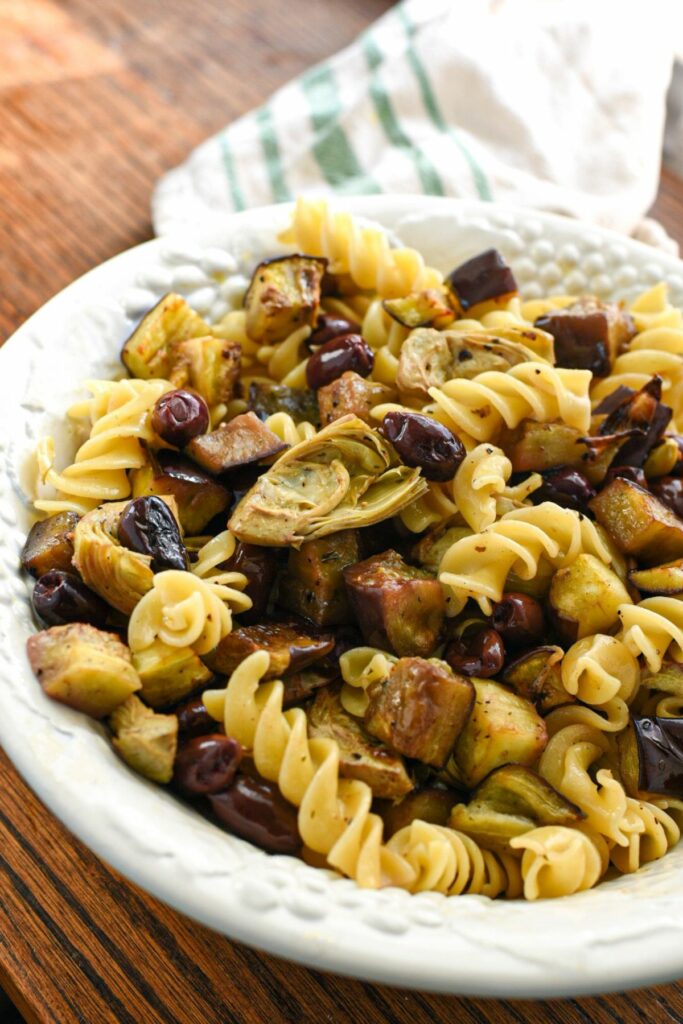 A large white porcelain serving bowl filled with fusilli pasta and roasted eggplant, olives and artichoke hearts
