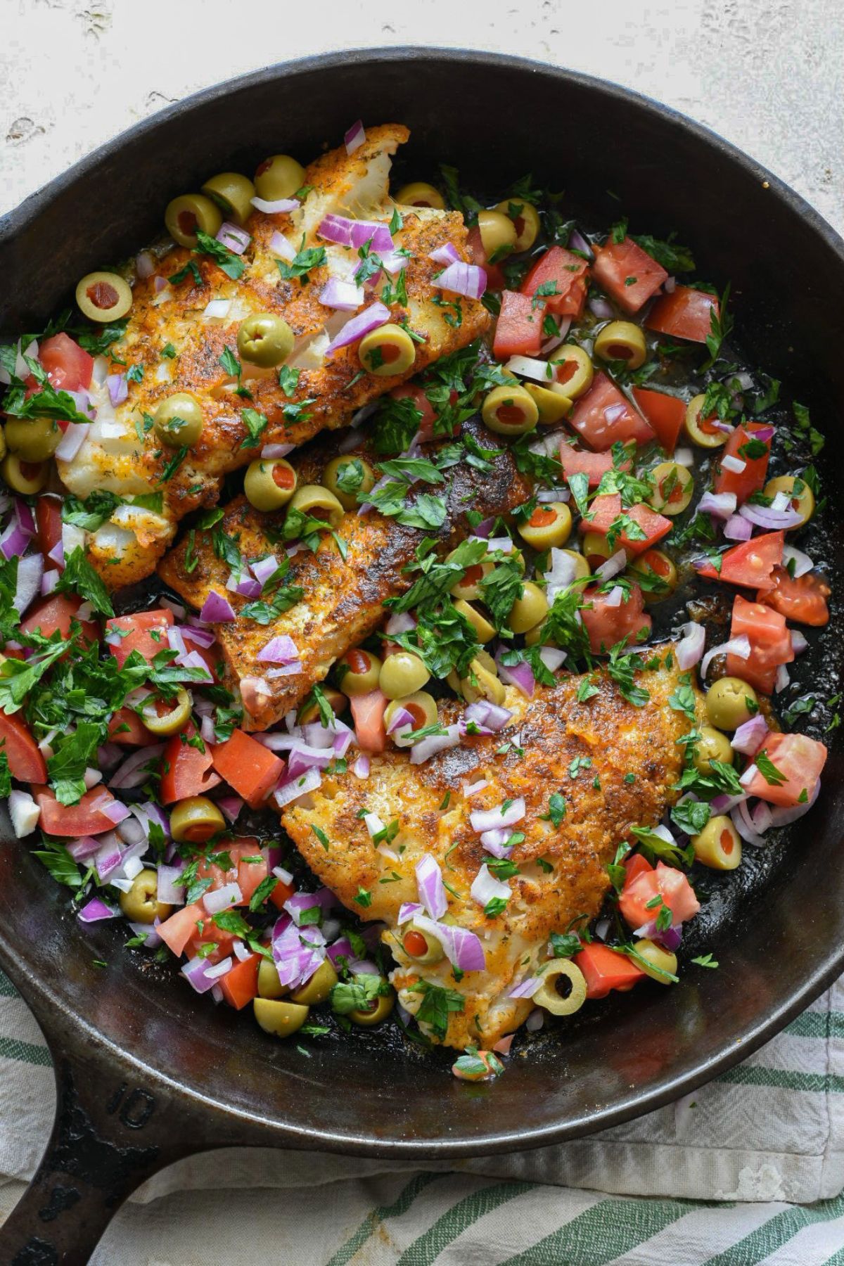 A cast iron skillet filled with three golden white fish fillets, green olives, chopped tomatoes, sliced red onion and green chopped parsley