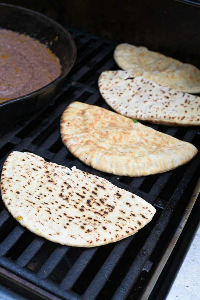 4 pita pockets on an outdoor gas fired grill being toasted, with a brown sauce in a cast iron skillet in the background