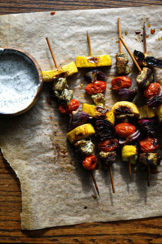 chicken and vegetable wooden skewers on parchment paper over a dark wood surface with a white sauce in a wooden bowl