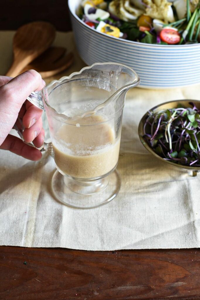 A clear glass gravy jar filled with caesar salad dressing, with a brass bowl of microgreens next to it and a large serving bowl of salad in the background