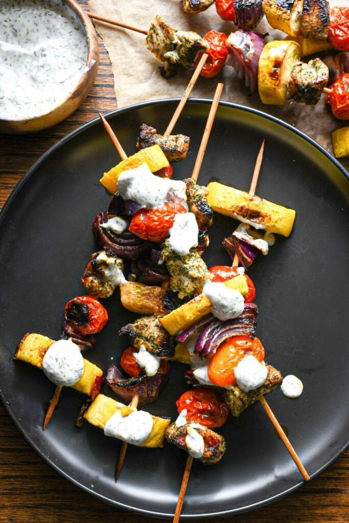 An overhead shot of a black plate filled with chicken and vegetable grilled kabobs, with a white greek yogurt sauce over the top and in the background.