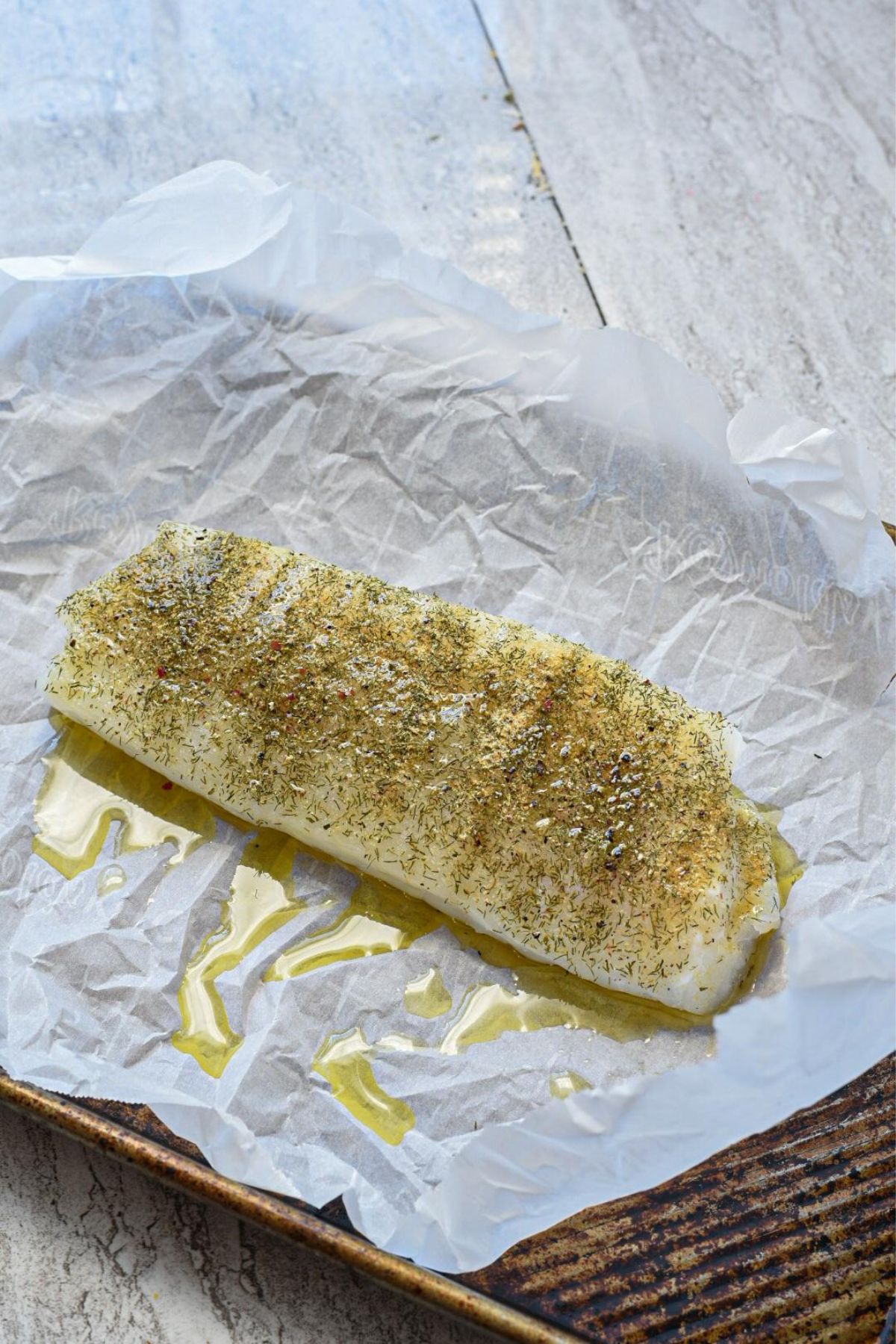 A piece of uncooked cod fish sprinkled with oil and dill on a parchment paper lined baking sheet