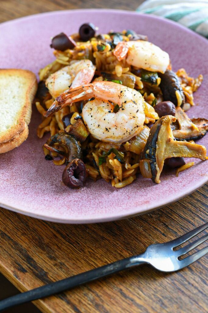 mediterranean shrimp orzo pasta and shiitake mushroom dinner on a pink plate with a piece of toasted baguette