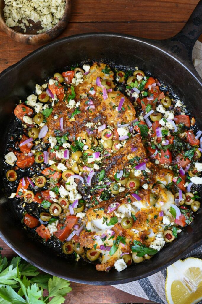 An overhead image of a large black cast iron skillet filled with three baked cod filets, topped with green olives, red onion, tomato, parsley and feta