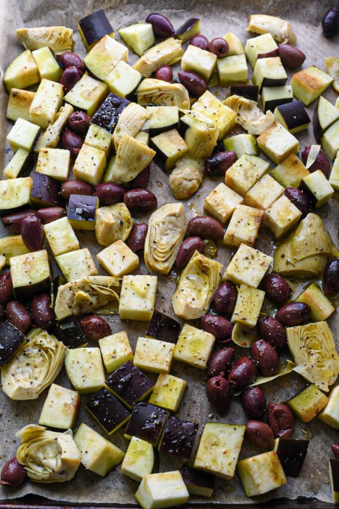 A sheet pan covered in parchment paper filled with cubed eggplant, kalamata olives and marinated artichoke hearts