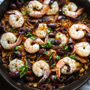 A cast iron skillet filled with mediterranean shrimp, olives, mushrooms and orzo, with green basil on top