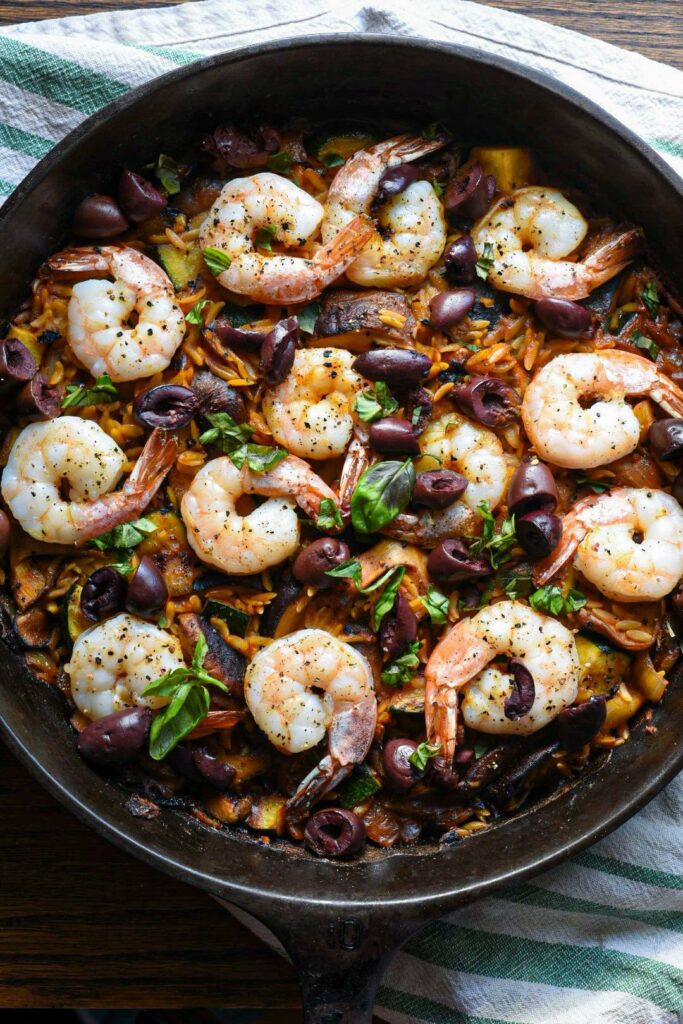 A cast iron skillet filled with mediterranean shrimp, olives, mushrooms and orzo, with green basil on top