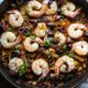 A cast iron skillet filled with mediterranean shrimp, olives, mushrooms and orzo, with green basil on top