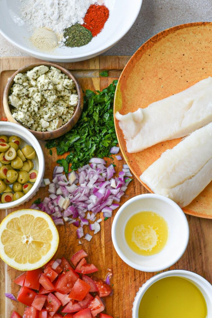 An overhead shot of the ingredients used to make this mediterranean cod recipe, including cod filets, butter, olive oil, chopped red onion and parsley, chopped tomatoes and olives, lemon, feta and flour with spices