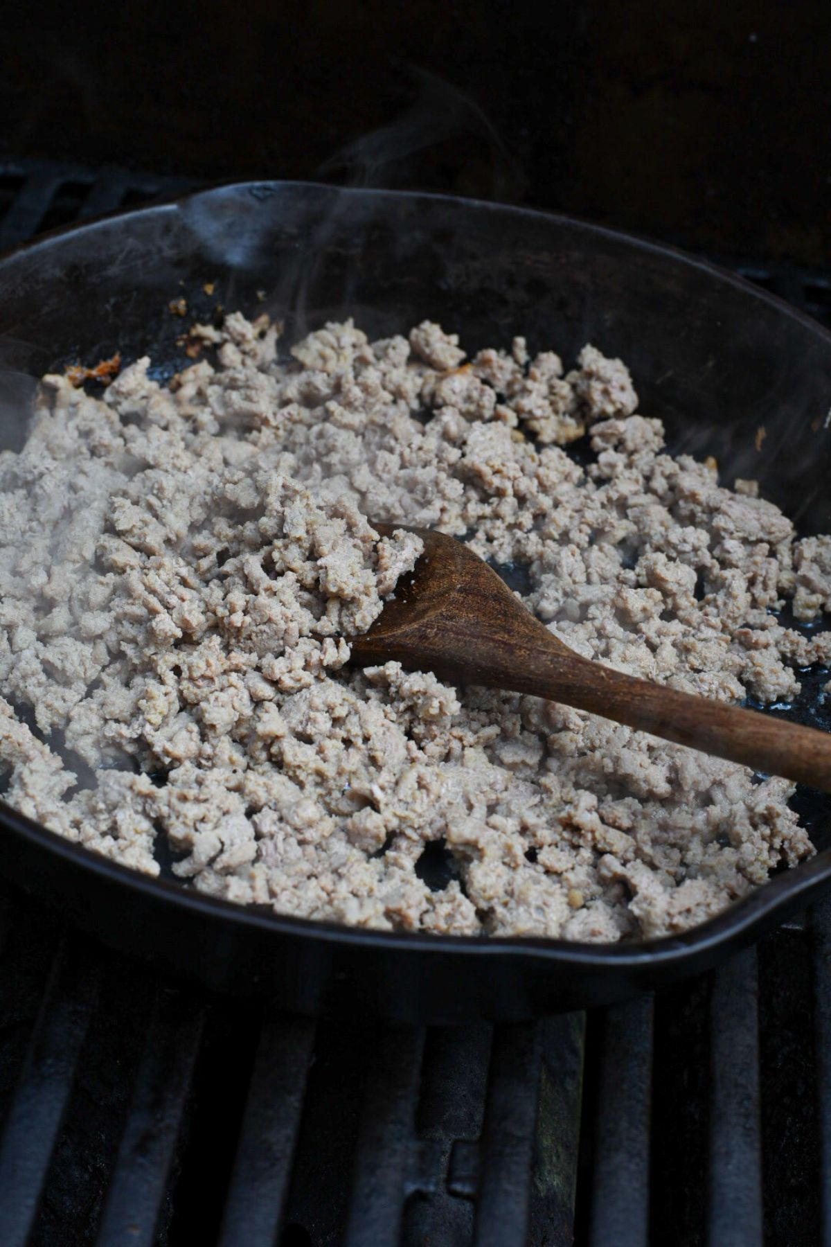 A cast iron skillet on a grill filled with ground turkey being stirred by a wooden spoon
