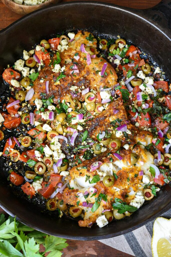 An overhead image of a large black cast iron skillet filled with three baked cod filets, topped with green olives, red onion, tomato, parsley and feta