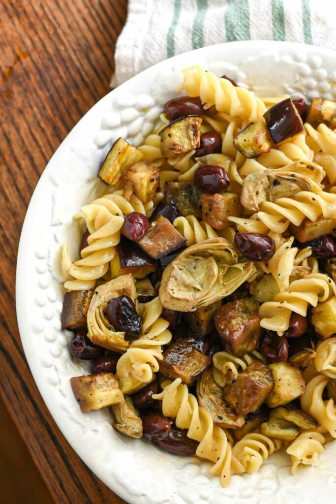 An overhead shot of a large white porcelain serving bowl filled with fusilli pasta and roasted eggplant, olives and artichoke hearts