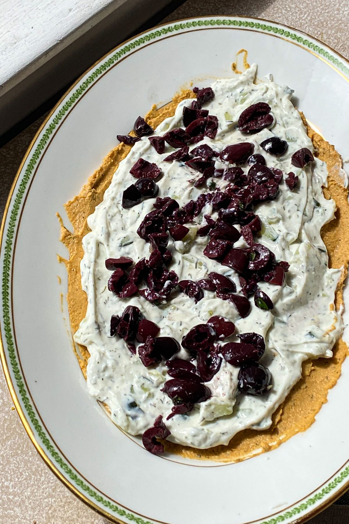 An oval white platter in the sun spread with a layer of hummus, a layer of tzatziki and chopped kalamata olives