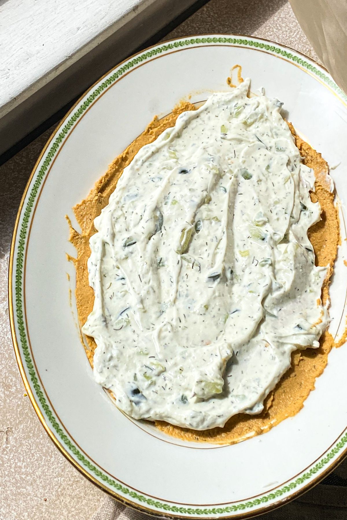 An oval white platter in the sun spread with a layer of hummus and a layer of tzatziki sauce