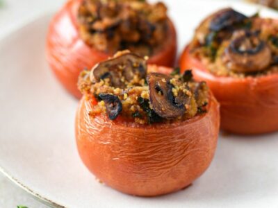 Three large beefsteak tomatoes that have been stuffed with mushrooms, kale and couscous on a white plate. The first tomato is in focus, while the others behind it are blurry.