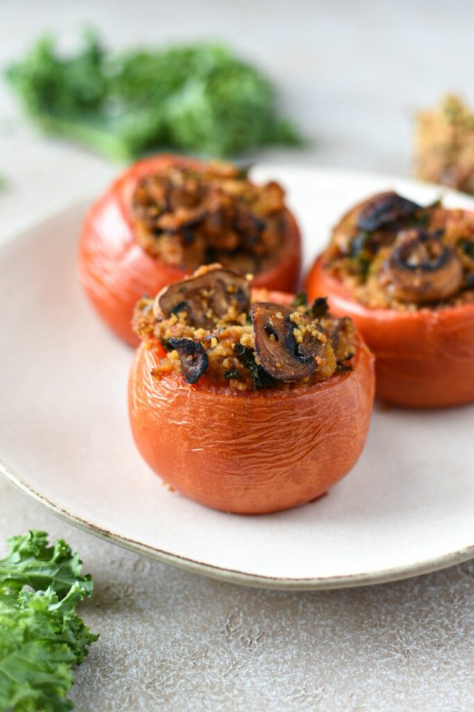 Three large beefsteak tomatoes that have been stuffed with mushrooms, kale and couscous on a white plate. The first tomato is in focus, while the others behind it are blurry.