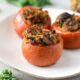 Three large beefsteak tomatoes that have been stuffed with mushrooms, kale and couscous on a white plate. The first tomato is in focus, while the others behind it are blurry.
