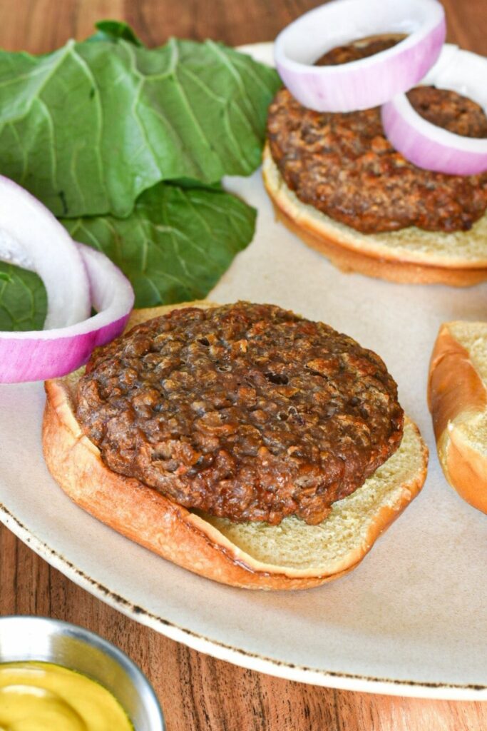 A beyond beef burger on a burger bun with red onion slices and greens behind them, and mustard in the corner of the shot