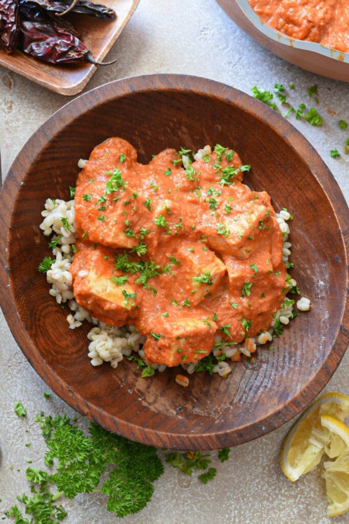 A wooden bowl filled with barley, tofu and arrabiata sauce with parsley sprinkled over the top and in the corner of the image.  There are serrano peppers in the top corner.