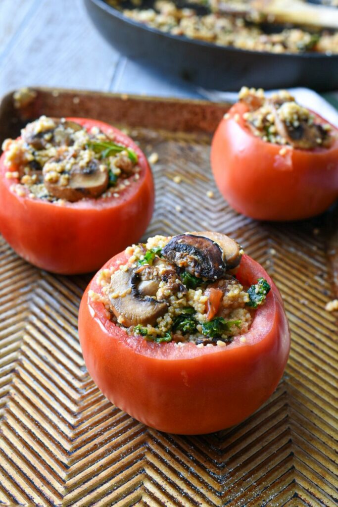 Three large beefsteak tomatoes on a ribbed baking sheet, filled with a mushroom couscous mixture in preparation for being baked