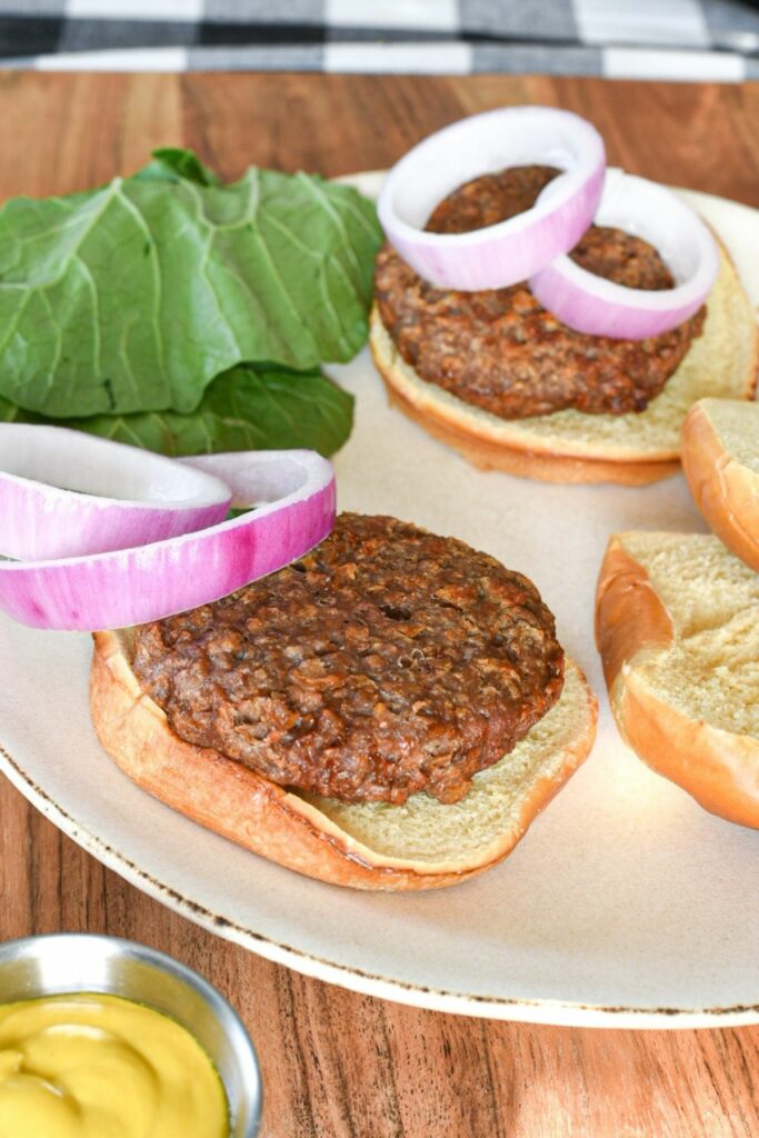 A white plate with a beyond beef burger on a burger bun with red onion slices and greens behind them, and mustard in the corner of the shot
