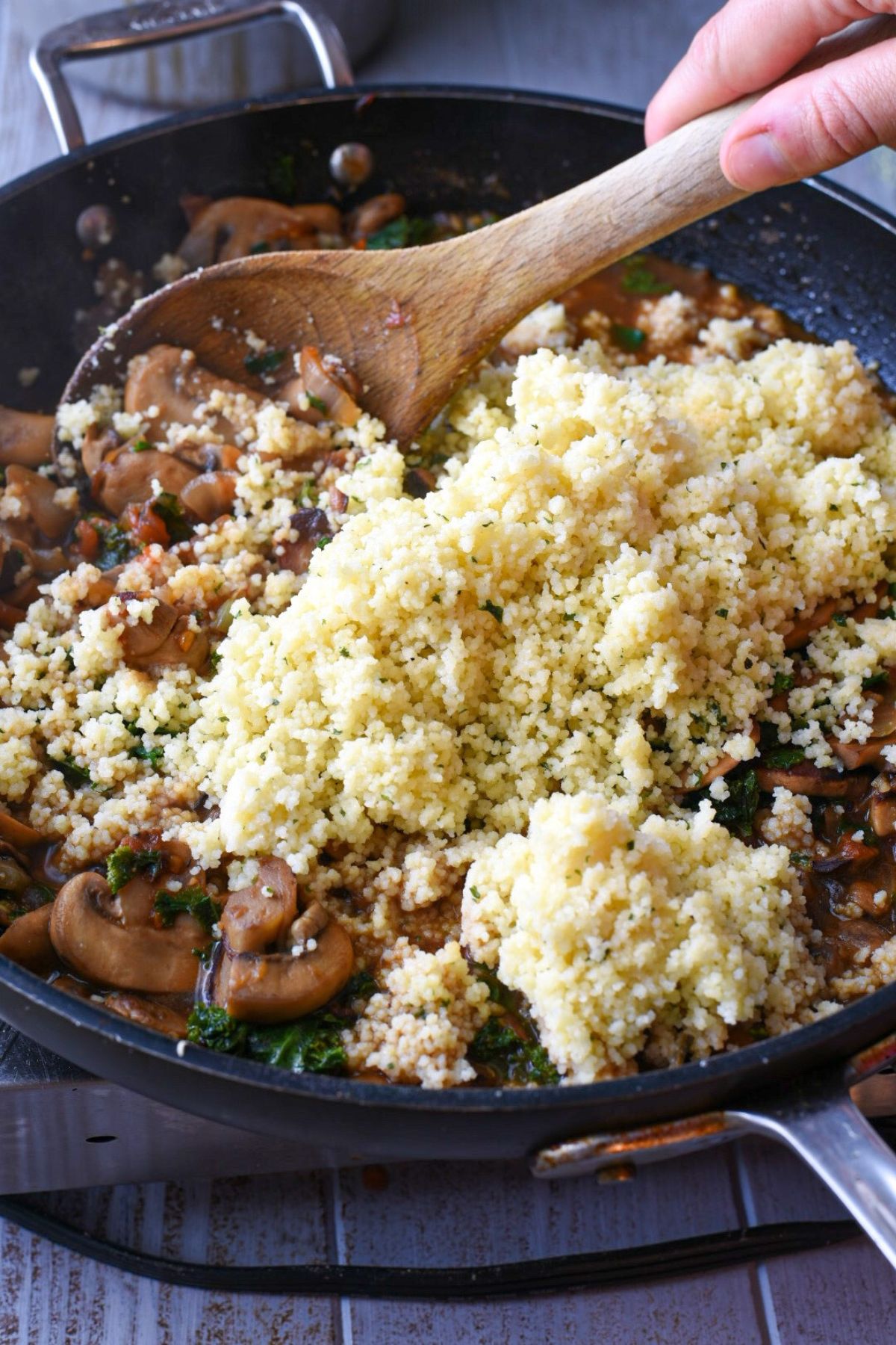 A nonstick pan filled with cooked mushrooms and kale, with cooked couscous poured over the top and a hand holding a wooden spoon mixing it