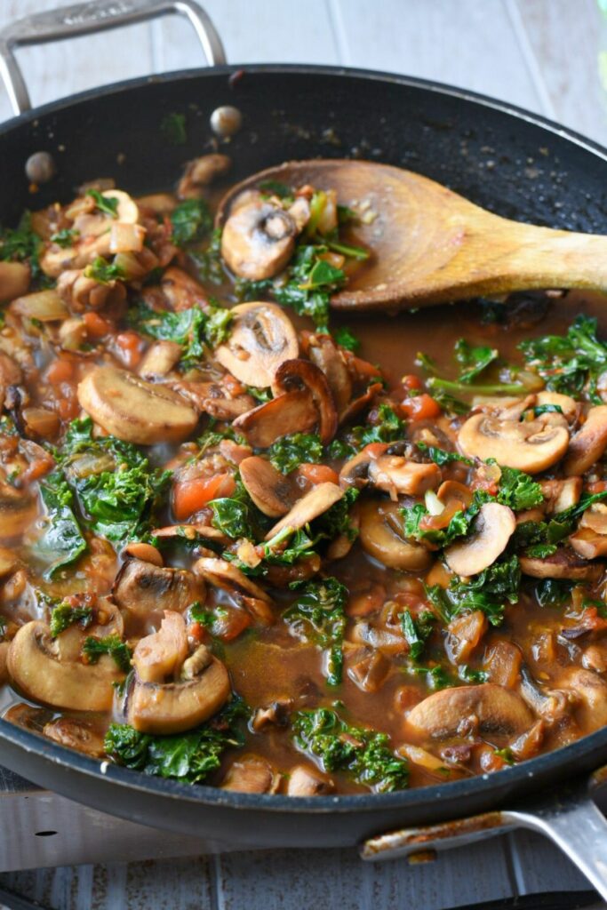 A large non-stick pan filled with cooked mushrooms, kale and tomatoes, with a brown broth around the vegetables and a wooden spoon in the pan