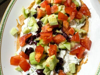 A plate filled with a greek layered dip with hummus, tzatziki, tomatoes, cucumber and olives